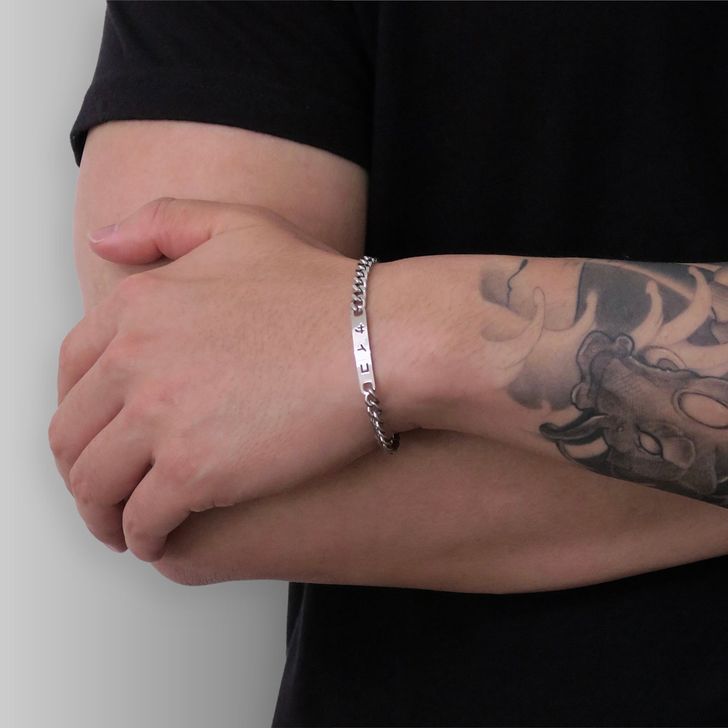 Male with tattooed arm crossing arms wearing 316L stainless steel cuban chain bracelet with Japanese SAIKO logo