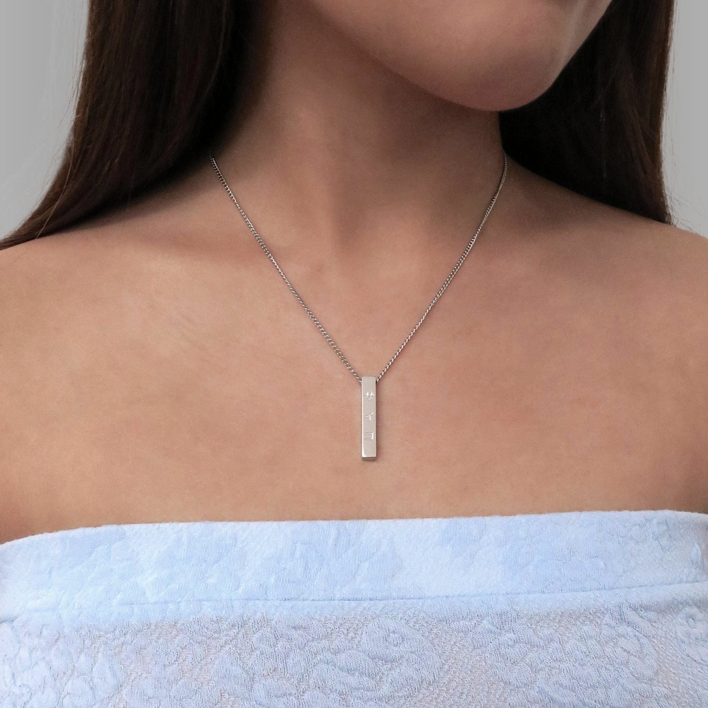 Girl with blue top wearing silver stainless steel 316L necklace with a vertical pendant and SAIKO logo hanging with a white background